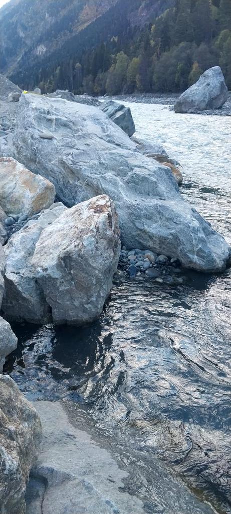 Крупные серые камни расположены вдоль берега реки. Вода в реке течет рядом с камнями, создавая небольшие волны и отражения на поверхности. На заднем плане видны лесистые горы, покрытые деревьями.