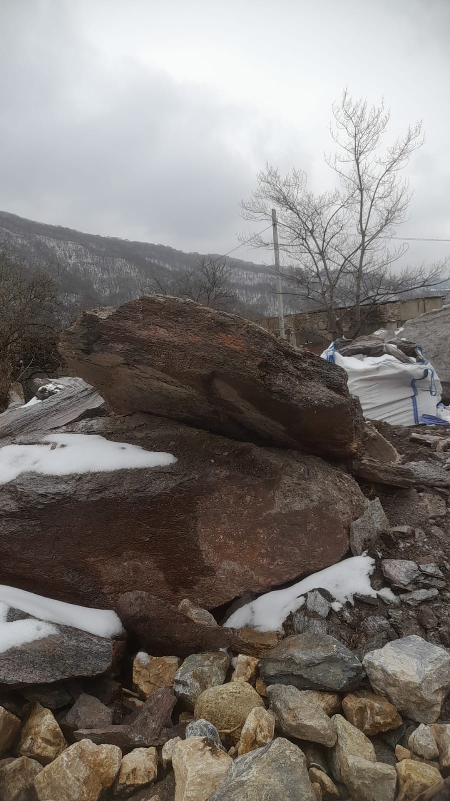 Крупные камни сланца серого, покрытые небольшим количеством снега. На заднем плане видны голые деревья и холмы, также покрытые снегом. Небо облачное и серое.