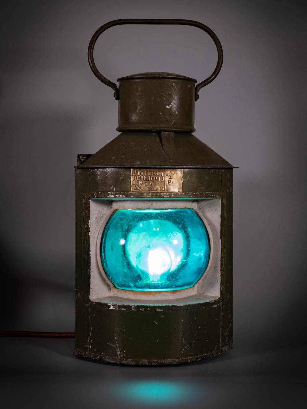 Навигационный судовой фонарь WOODandROOT 1957 года с холодным сине-зелёным свечением, подчёркивающим глубину стекла и старого корпуса.