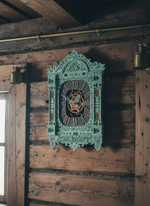 Мятные резные часы-наличник WOODandROOT висят на деревянной стене, тёплый свет подчёркивает северный характер резьбы и многослойной покраски.