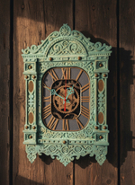 Мятные часы-наличник WOODandROOT на деревянной стене, мягкий тёплый свет выделяет резные элементы и многослойную окраску.