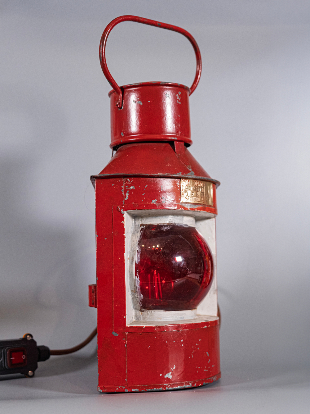Винтажный корабельный фонарь WOODandROOT 1957 года в красном корпусе с рубиновым стеклом, стоящий на столе с тканевым проводом и выключателем.