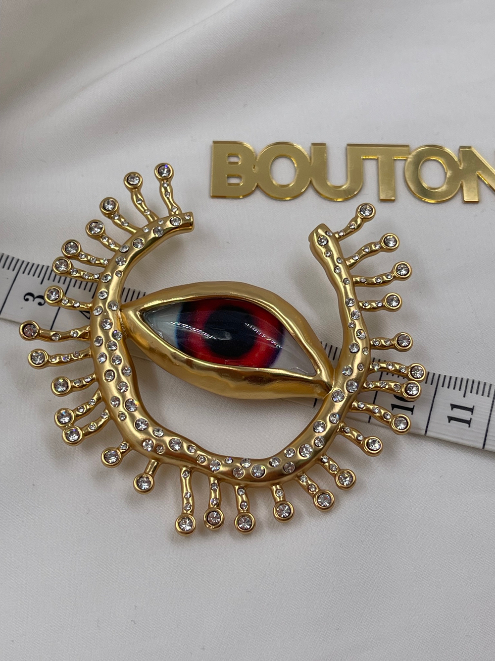Брошь Скиап. золотистая в форме глаза с подковой из ресниц со стразами красный