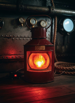 Винтажный корабельный фонарь WOODandROOT 1957 года с тёплым красным светом, стоящий в тёмном судовом интерьере с металлическими приборами и канатами.