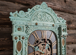 Верхняя часть мятных резных часов-наличника WOODandROOT на фоне старого деревянного сруба, слоистая покраска подчёркивает глубину северного орнамента.