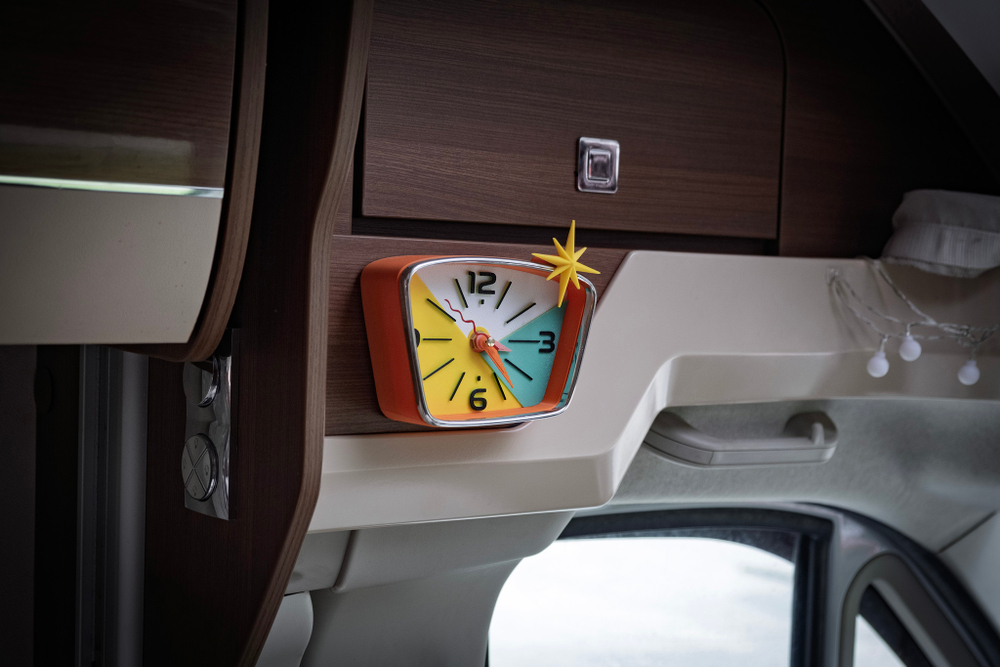 Настенные мини‑часы Camper Starburst 15×12 см с оранжевым корпусом и звездой‑компасом, прикреплены к деревянной панели автодома, создают уют и яркий ретро стиль.