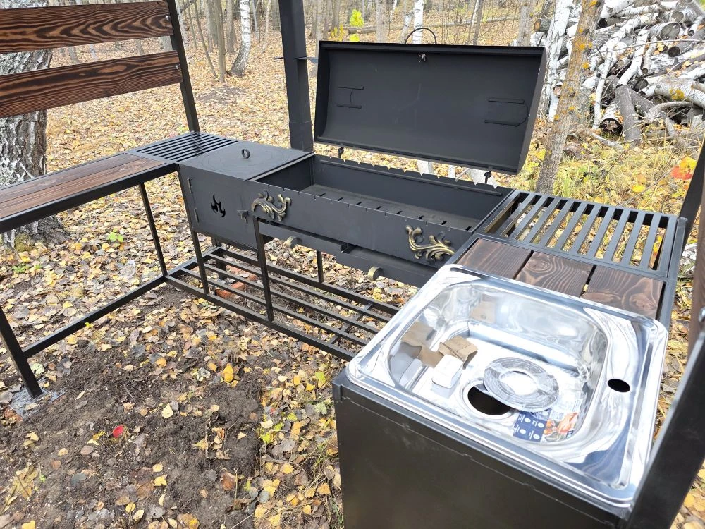 Мангальная зона из стали 5 мм с мойкой (Казань) Если вы хотите купить мангальную зону с мойкой в Казани, присмотритесь к нашему варианту: это мангальная зона 5 мм с крышей (полноценная уличная кухня/зона барбекю), где есть мангал 5 мм с жаровней 90×40×15 (рассчитан примерно на 11 шампуров), крышка барбекю с термодатчиком и шиберами для удобной регулировки тяги и температуры. Также зону можно использовать как коптильню — коптить рыбу, мясо и курицу.  Отдельно предусмотрена печь под казан 5 мм размером 40×40×30 с кольцами под казаны 3/4/6/8/10/12 литров, есть труба 80×80 с заслонкой, колосник и зольник. Для удобства сделаны столик для нарезки (стол металл+дерево), нержавеющая мойка 48×48, дровница и тубус для шампуров. Конструкция разборная, собирается на болтовых соединениях. Зона может быть в наличии в Казани, доступна доставка и установка, а также изготовление мангальной зоны под заказ под ваши размеры.